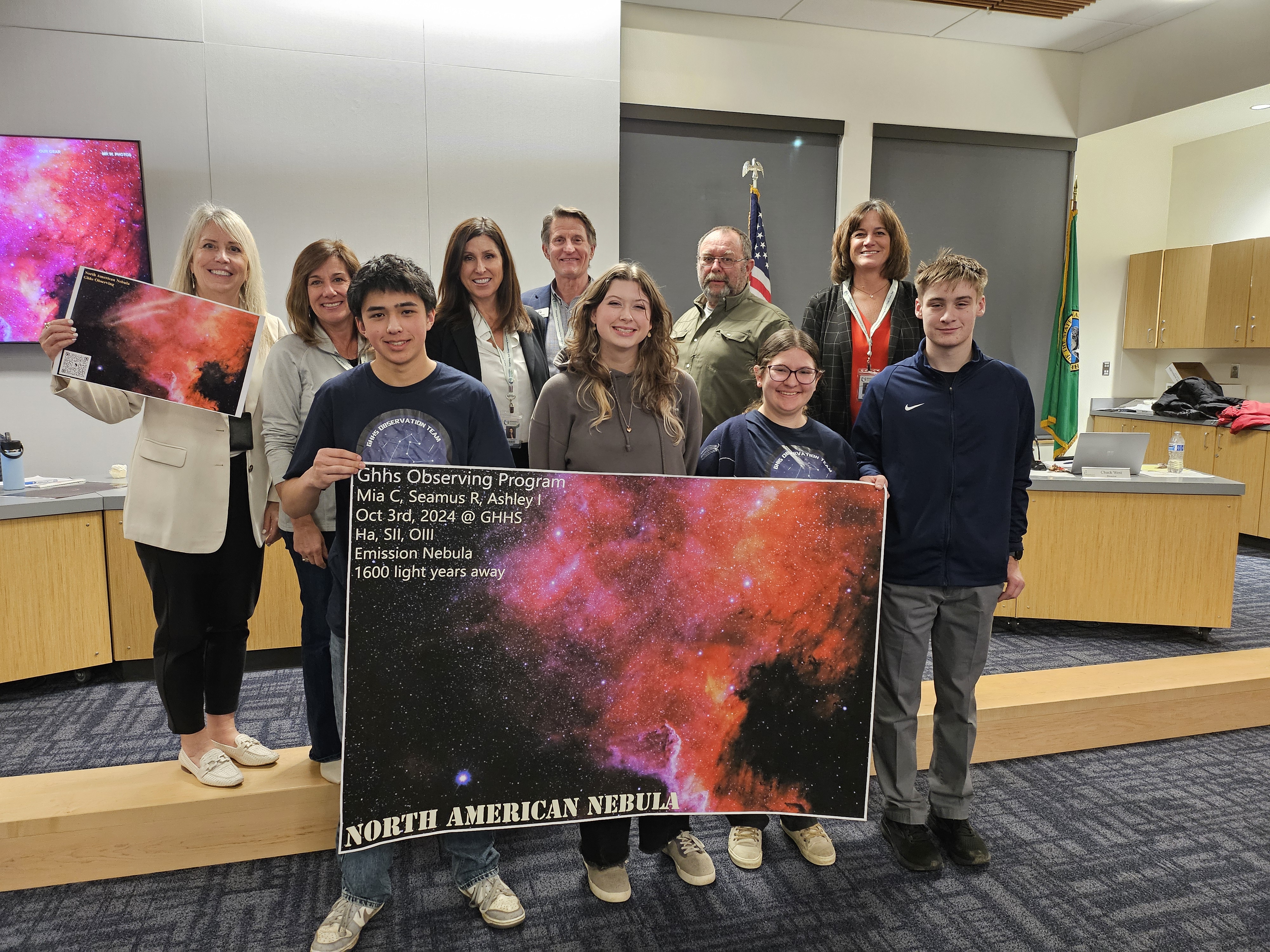 A group photo from a school board meeting showing students from the Gig Harbor High School Observing Program alongside board members and administrators. The students are proudly displaying large photographs of the North American Nebula they captured, showing vibrant red and pink cosmic clouds against a dark space background. The image includes a mounted display on the wall behind them showing similar deep space photography. The group stands in front of an American flag and Washington state flag in what appears to be a school district board room.