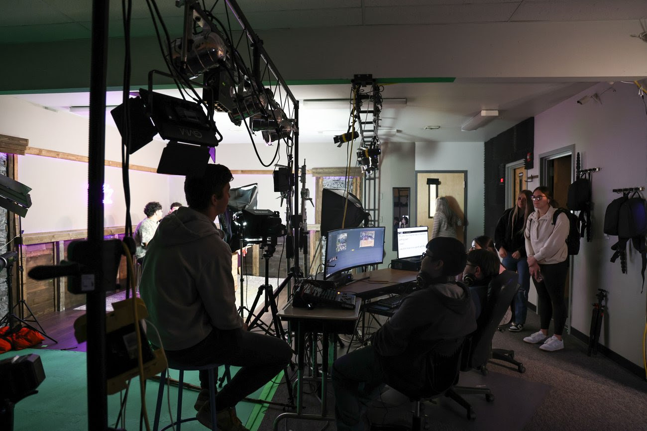 A group of young people work on a video production in a studio setting, with cameras, lights, and computer monitors.