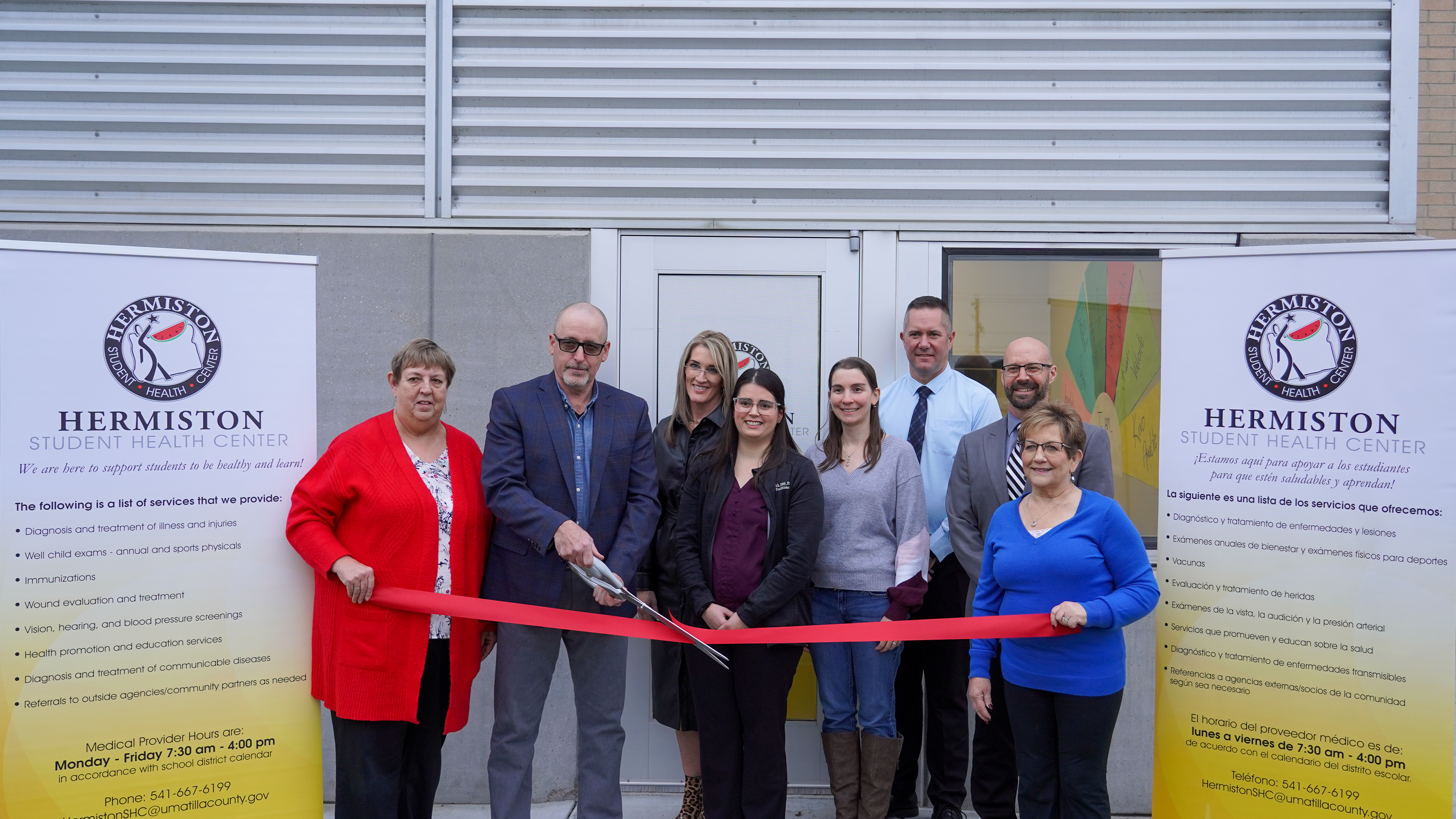 A group of people stand in front of a building, holding a red ribbon and scissors, celebrating the opening of the Hermiston Student Health Center.