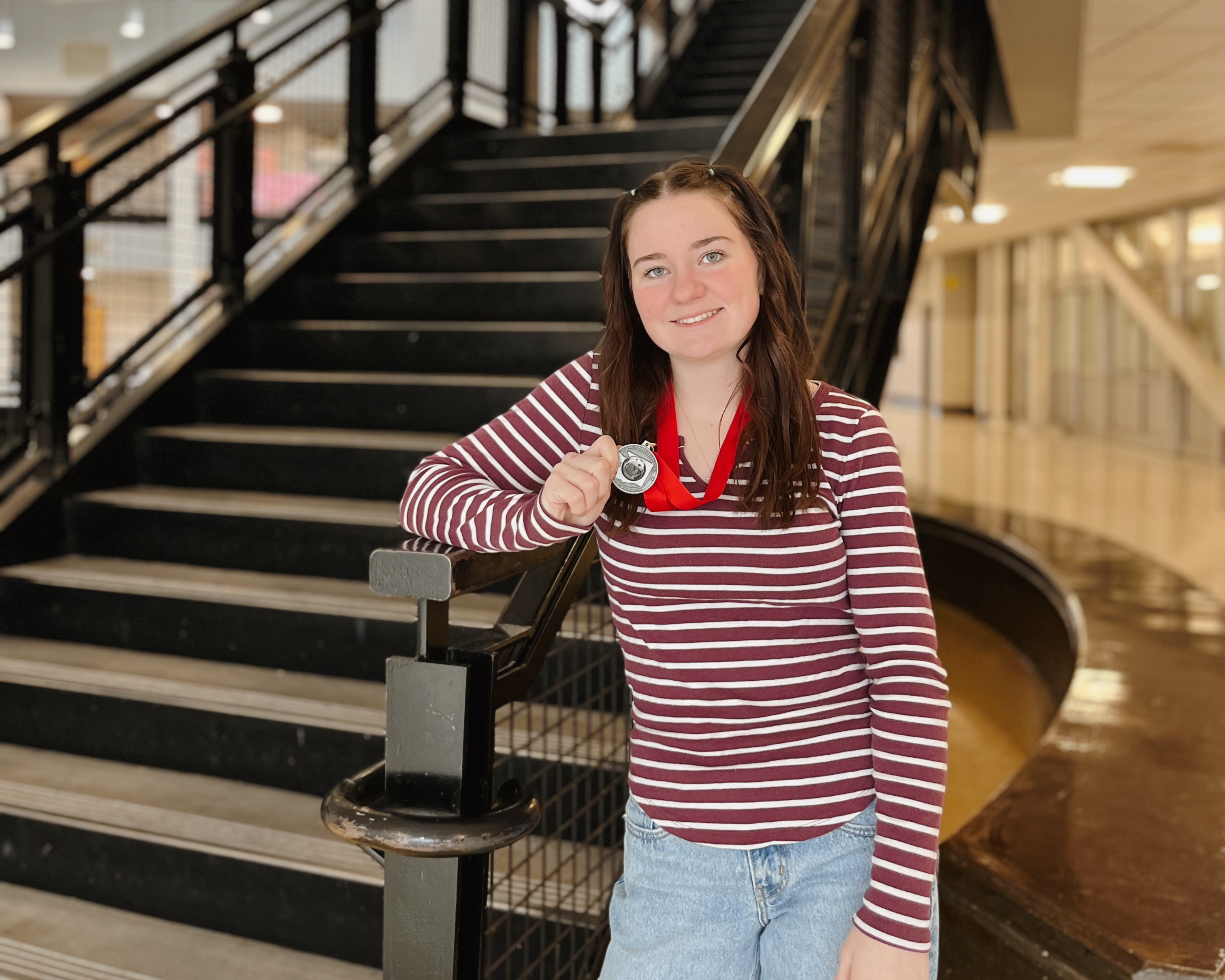 A young woman in a striped shirt smiles while holding a medal, standing on a staircase.