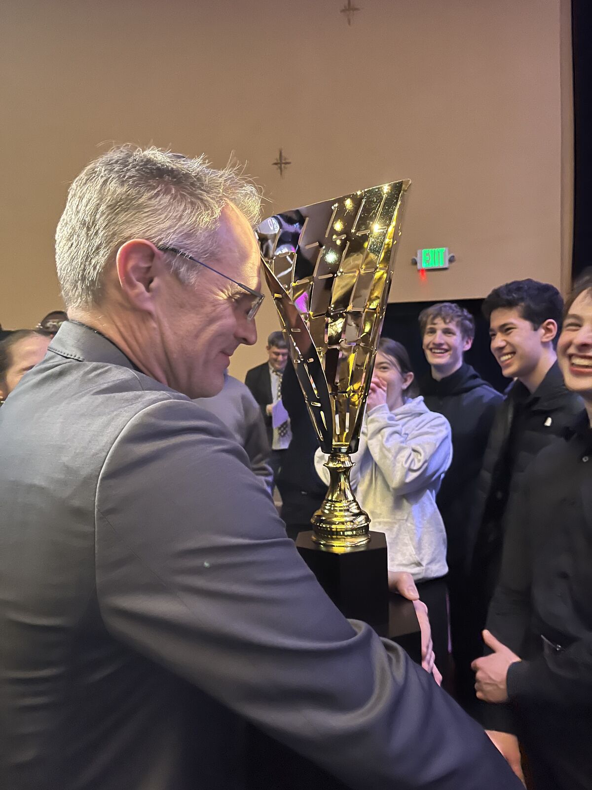 A man in a suit holds a large gold trophy while surrounded by a group of smiling young people.