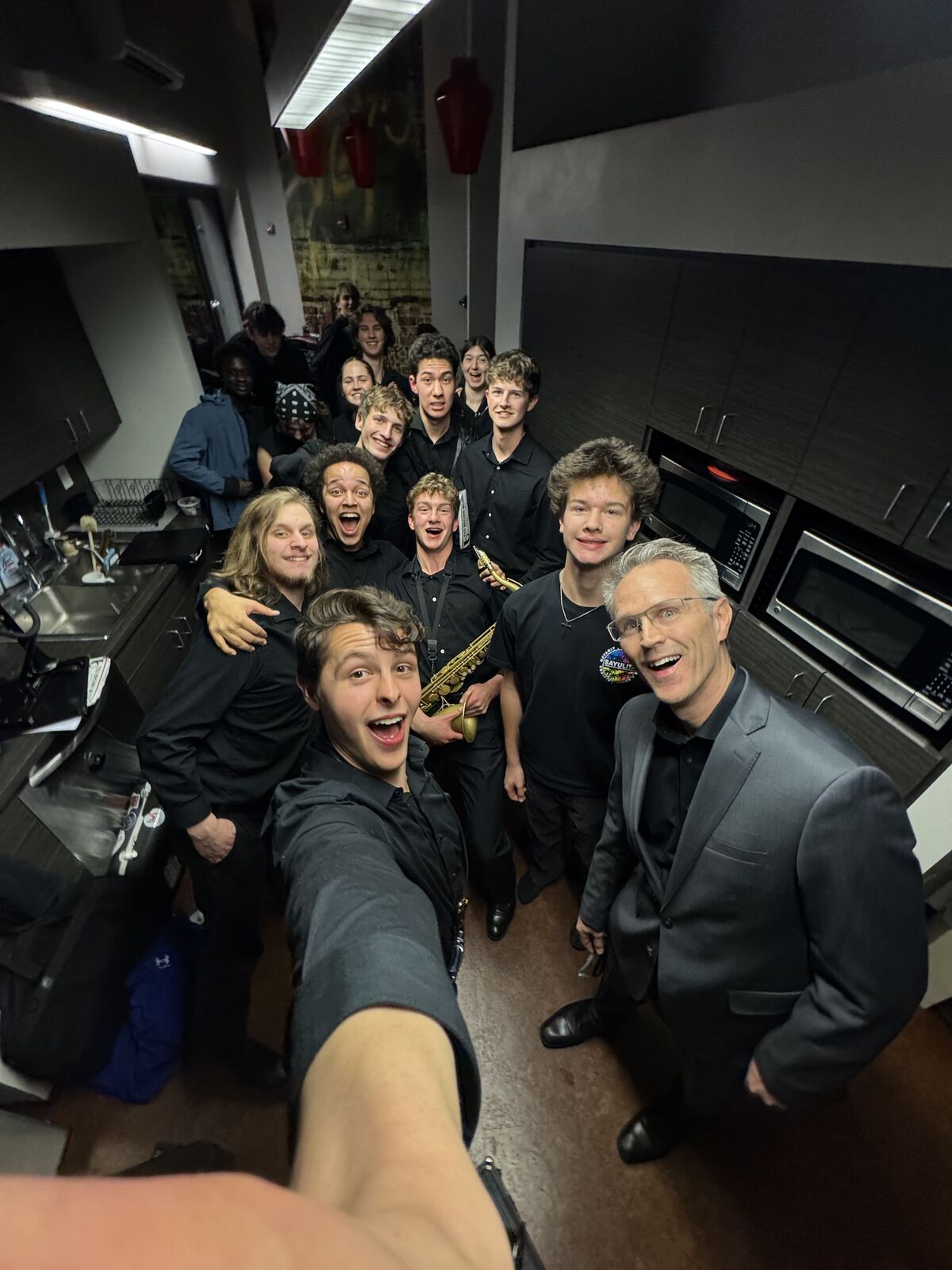 A group of musicians pose for a selfie in a backstage area, with a saxophone visible.