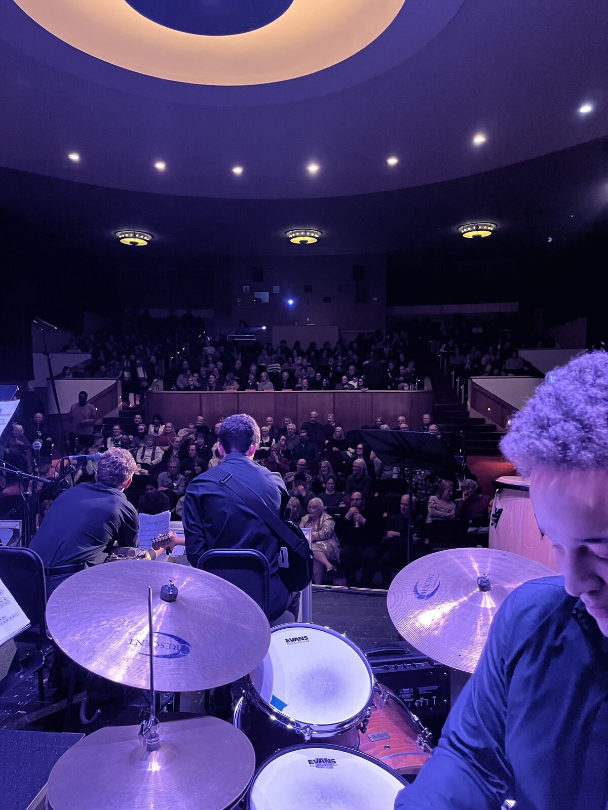 A drummer performs on stage in front of a large audience in a theater.