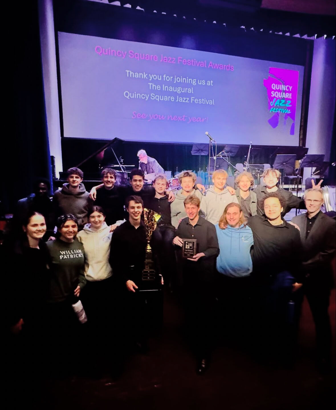 A group of musicians pose for a photo after winning awards at the Quincy Square Jazz Festival. The sign reads 'Thank you for joining us at The Inaugural Quincy Square Jazz Festival. See you next year!'