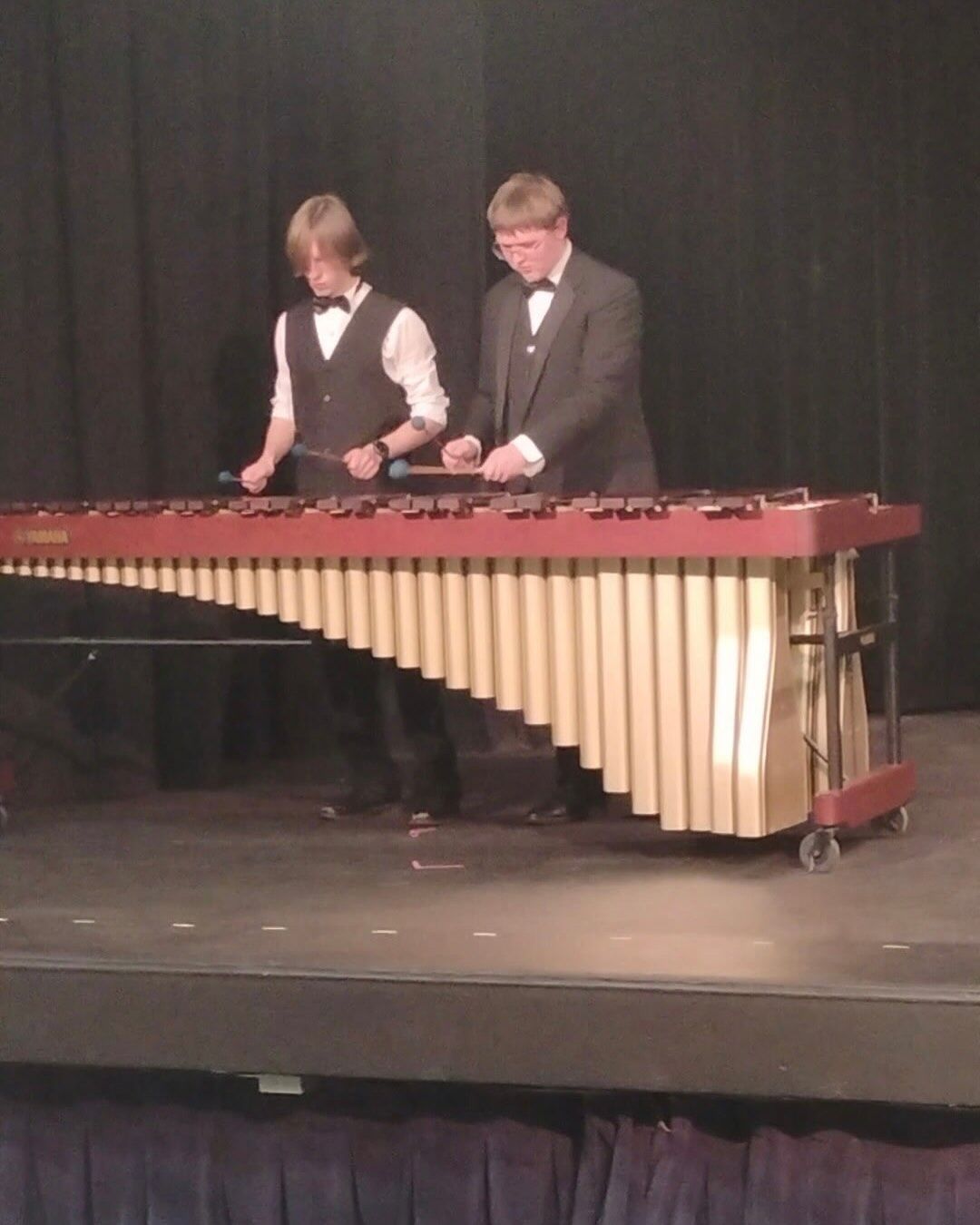 Two young men play a marimba on a stage, one in a suit and the other in a vest and shirt.