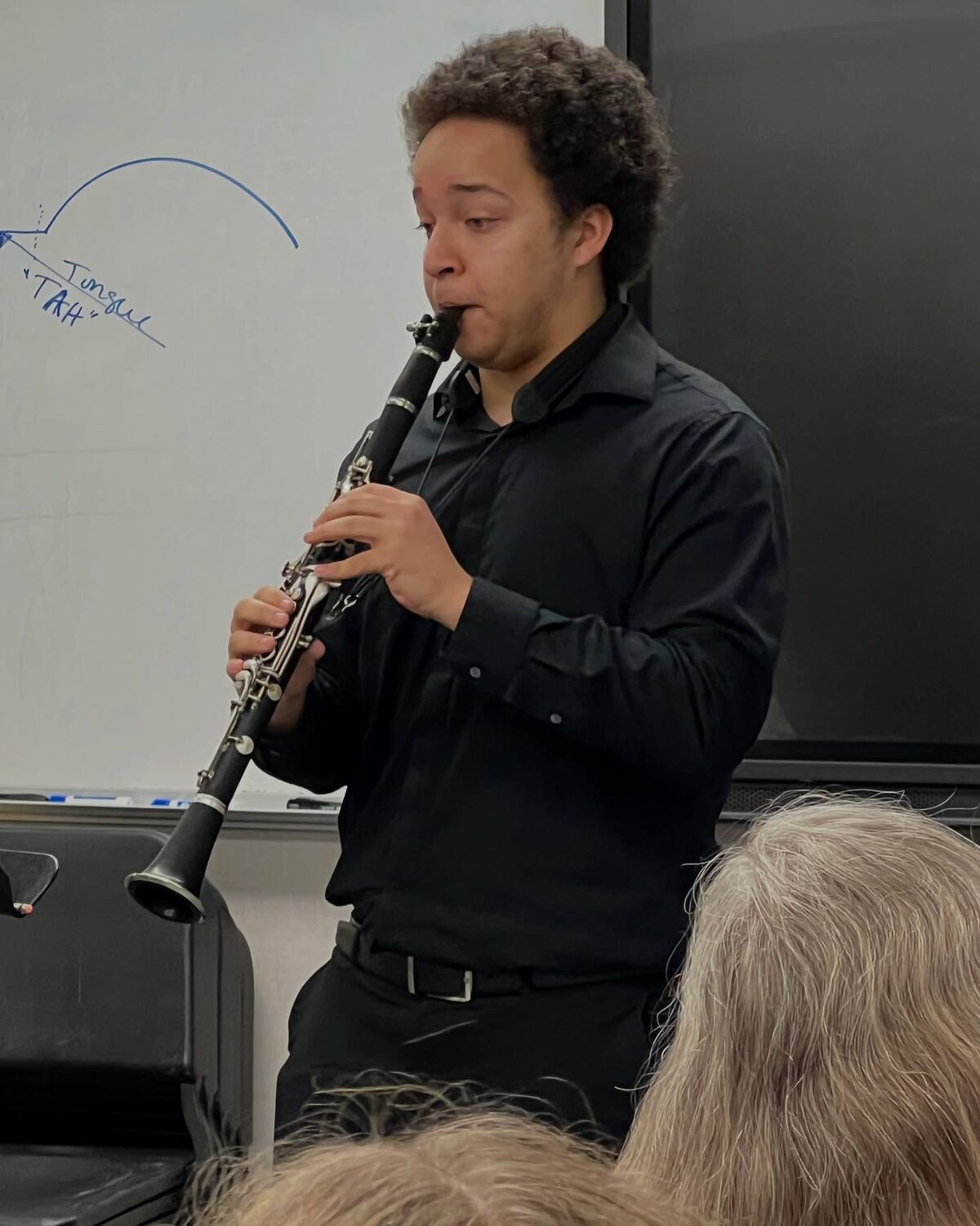 A young man plays the clarinet during a performance, with a whiteboard and audience members visible in the background.