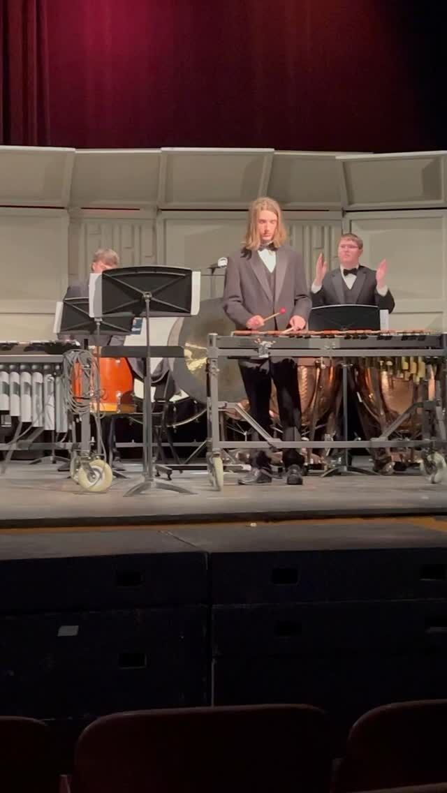 A young man plays the xylophone on stage during a performance, with other musicians behind him.