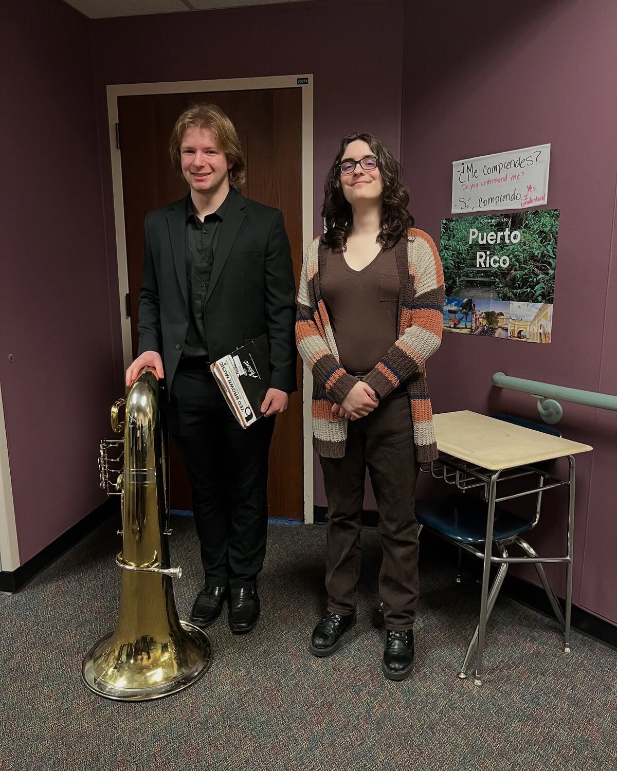 Two young musicians stand in a hallway, one holding a large tuba and the other a music book. A poster for Puerto Rico is visible on the wall.