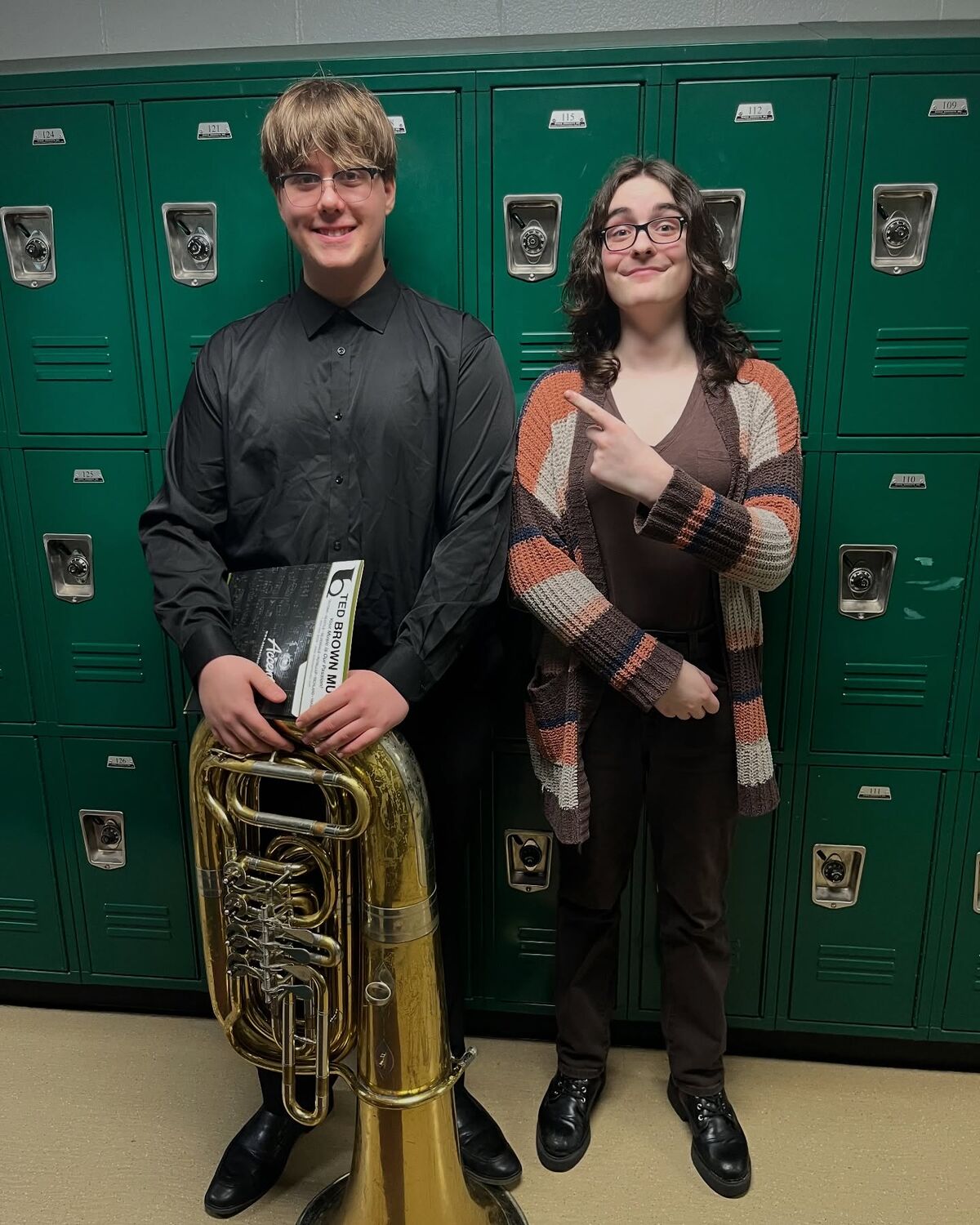 Two students stand in front of a row of green lockers, one holding a large tuba.