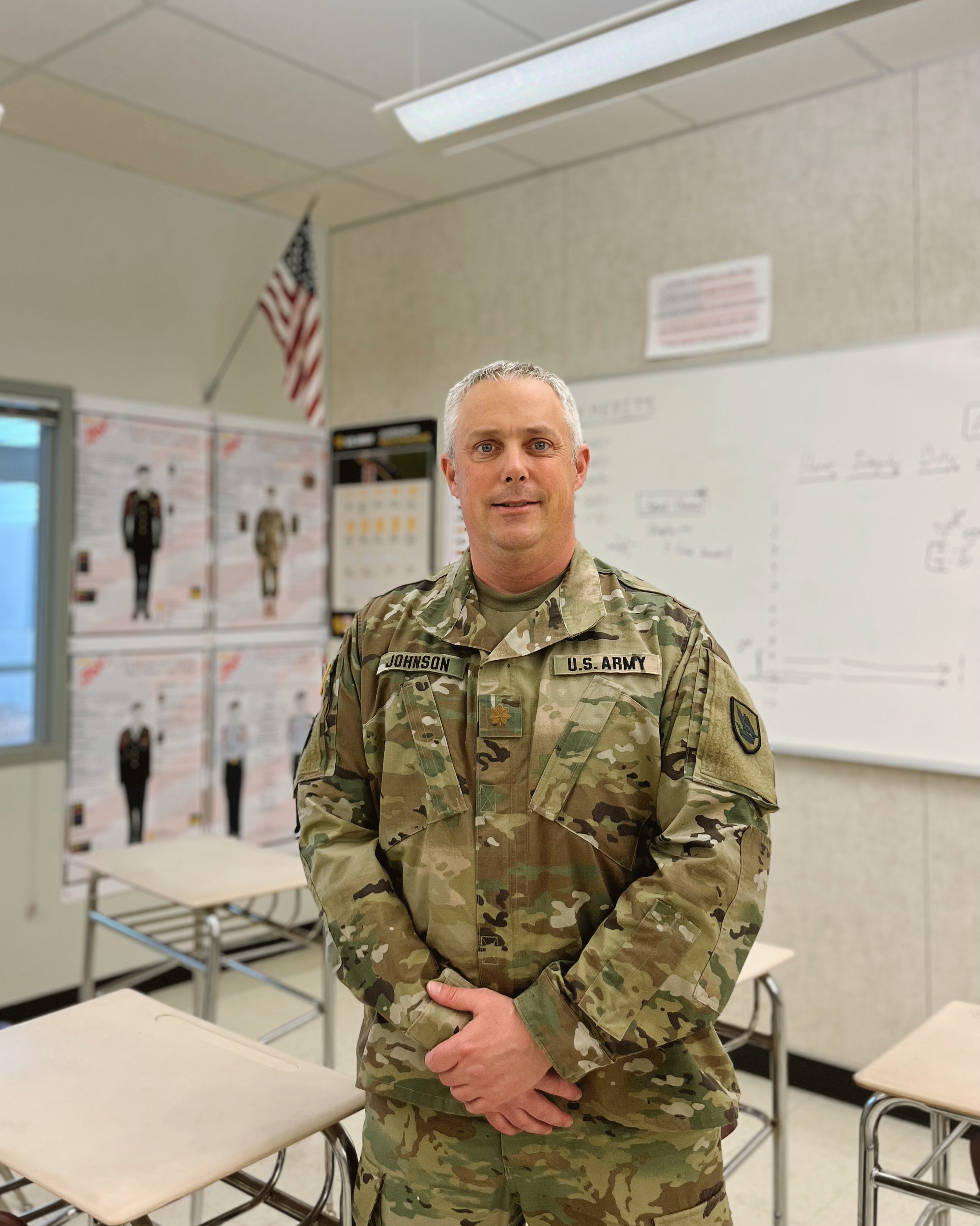 A man in a U.S. Army uniform stands in a classroom with a whiteboard and desks.