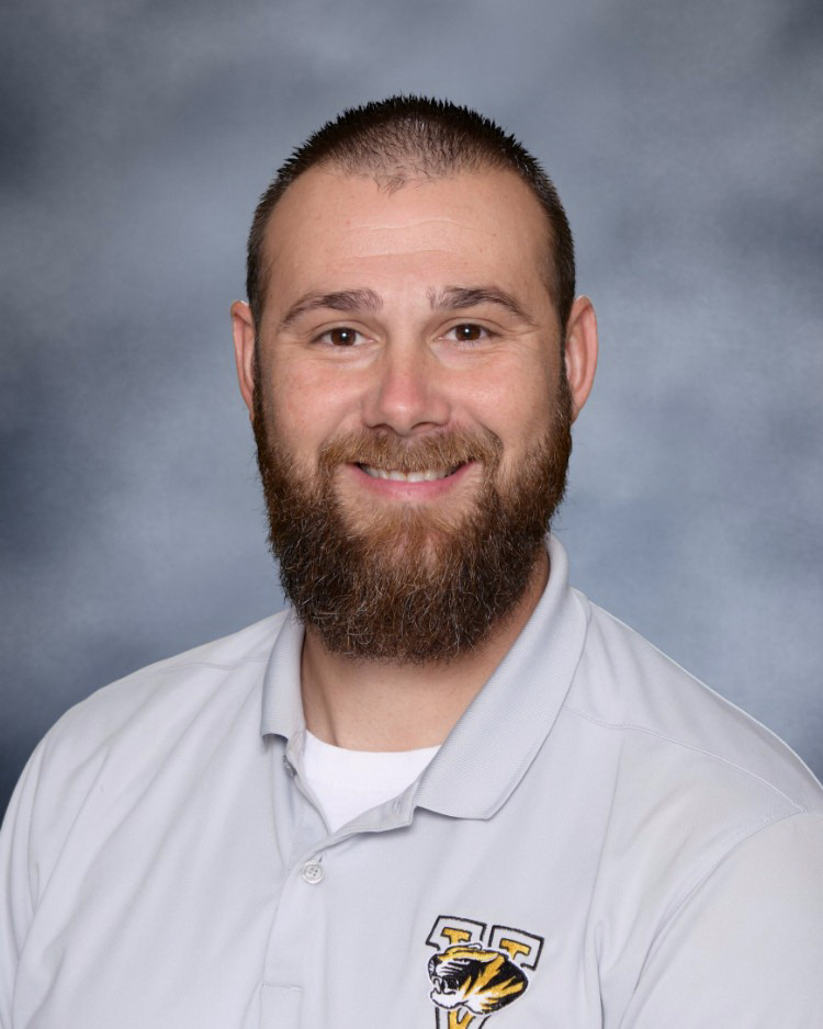 A man with a beard smiles for a portrait, wearing a light gray polo shirt with a tiger logo.