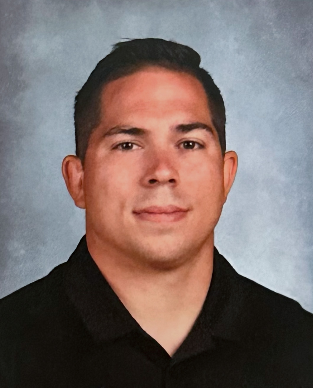 A headshot of a man with short brown hair wearing a black collared shirt against a light blue background.