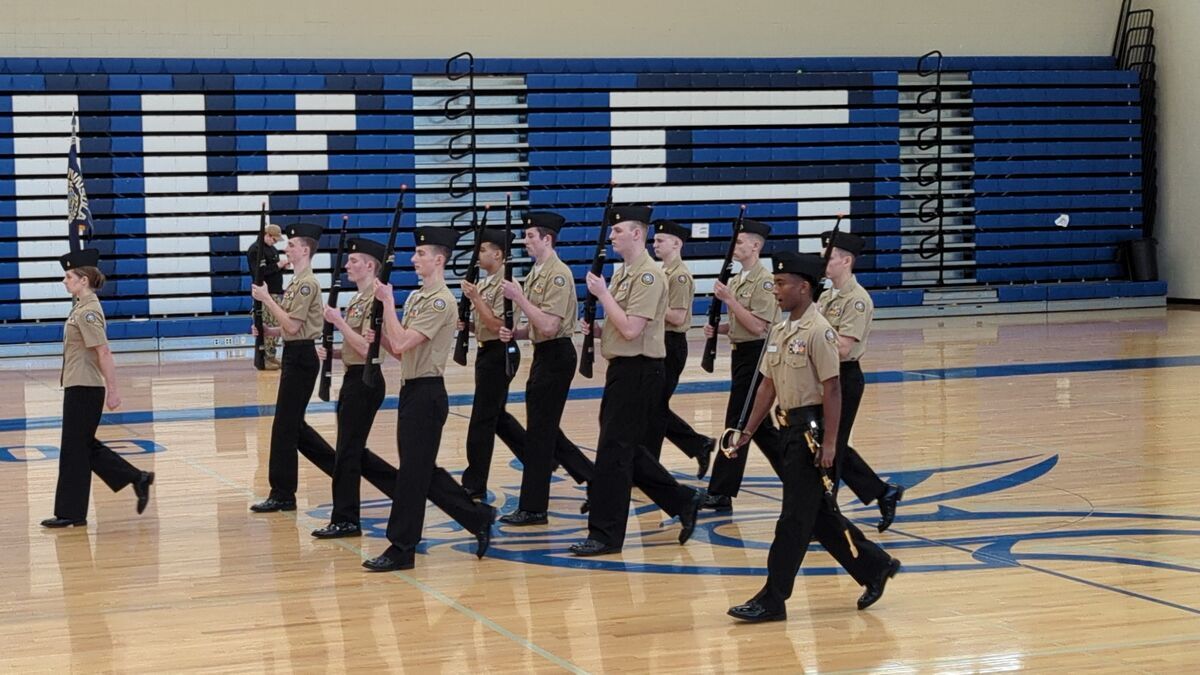 A group of young people in uniform march in a gymnasium, holding rifles.