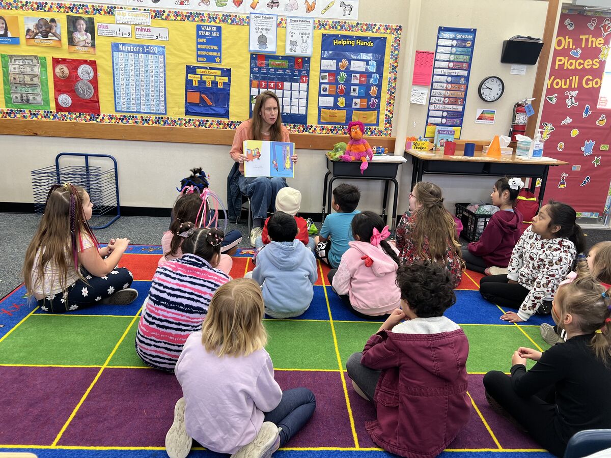 A teacher reads a story to a group of students sitting on a colorful rug in a classroom.