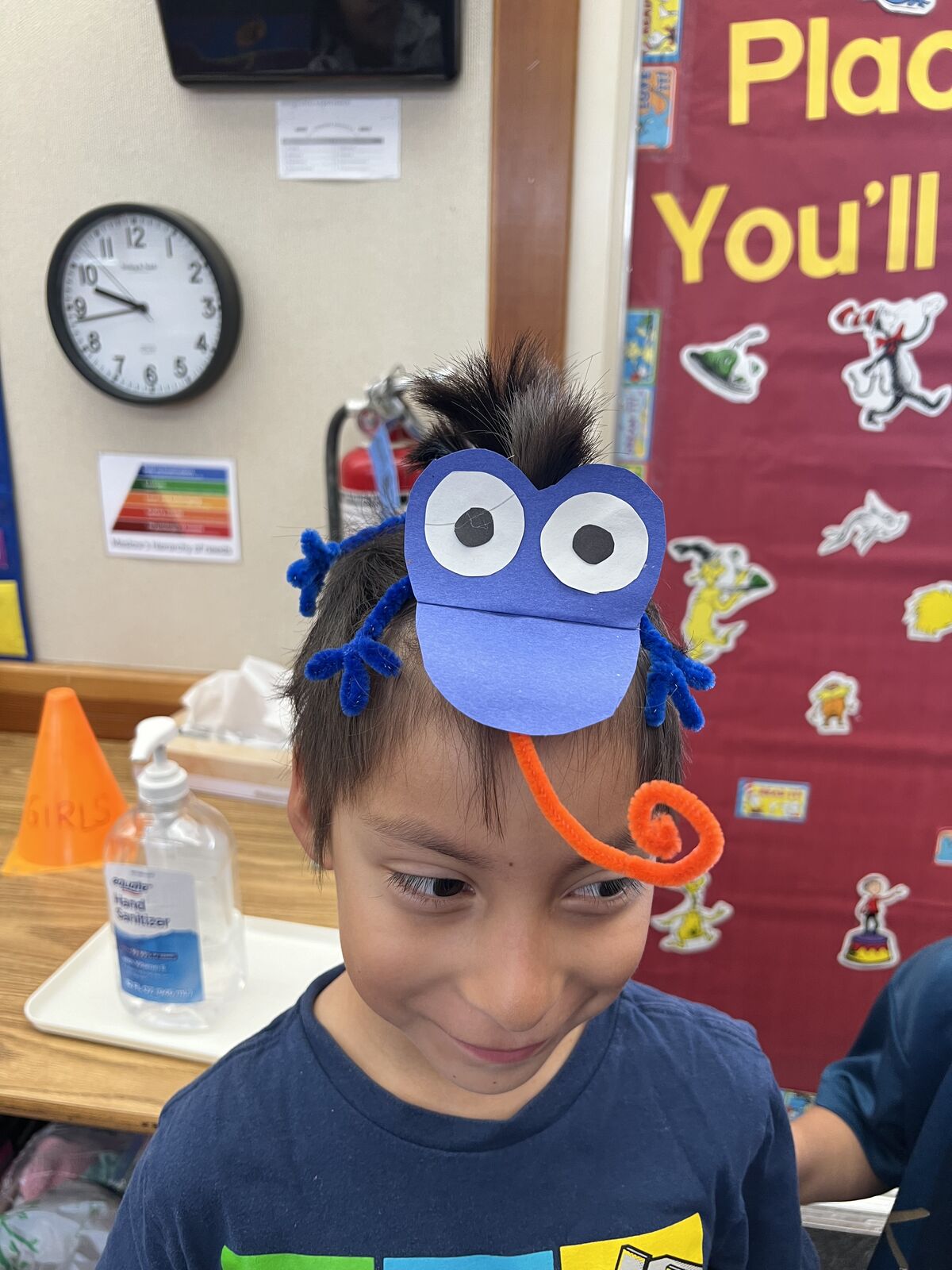 A young student wears a colorful frog headband in a classroom setting.