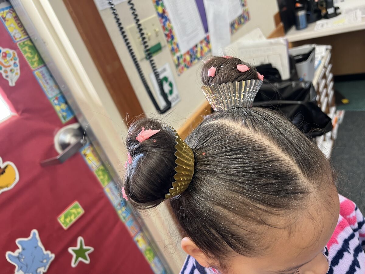 A child's hair is styled with two buns, each topped with a cupcake liner and pink frosting.