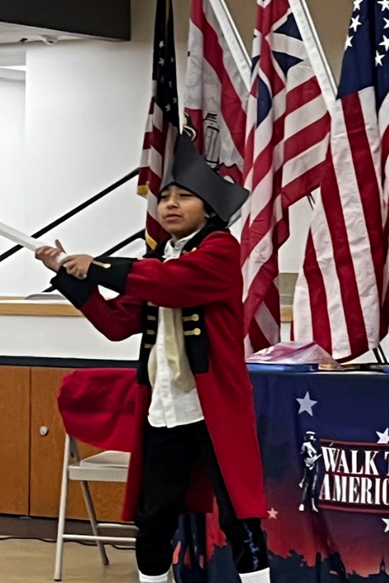 Image of a 5th-grade student passionately stepping into the shoes of an iconic historical figure, dressed in a colonial-era costume complete with a red coat, gold-trimmed black lapels, and a tricorn hat. 