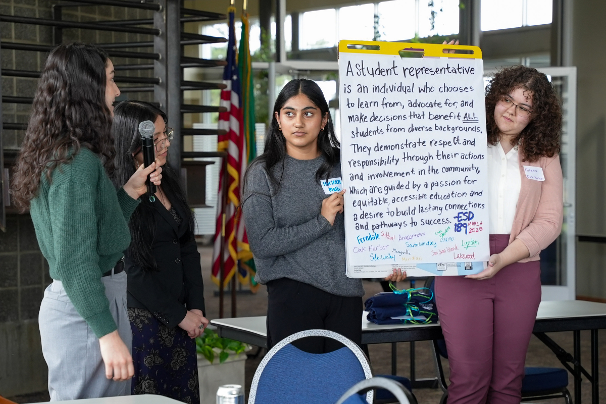 4 students present a handwritten mission statement that two of them are holding up while another speaks into a microphone