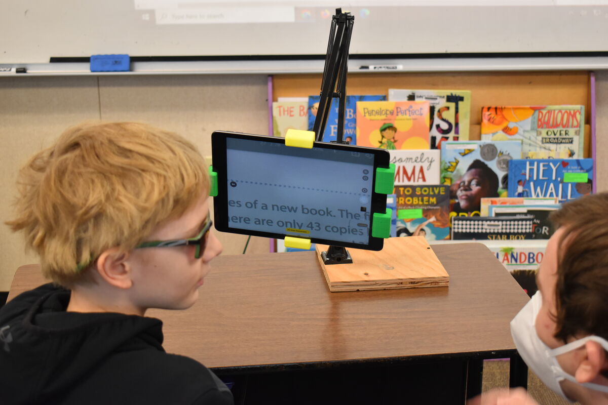 A student wearing glasses looks at a tablet displaying text, while another student sits behind them wearing a face mask.