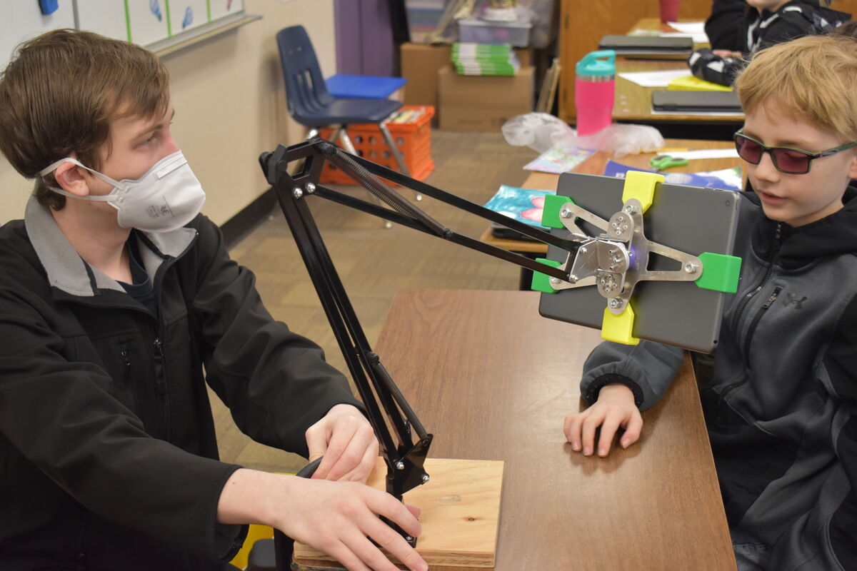 A young boy wearing glasses looks at a tablet held by a flexible arm, while a person wearing a face mask adjusts the arm.