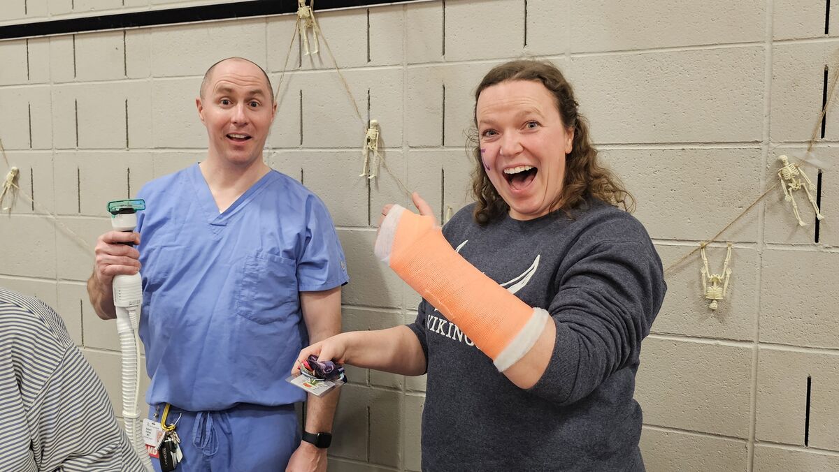 Two people stand in front of a brick wall decorated with hanging skeletons. The person on the left is holding a medical device, while the person on the right has a cast on their arm and is pointing with their other hand.