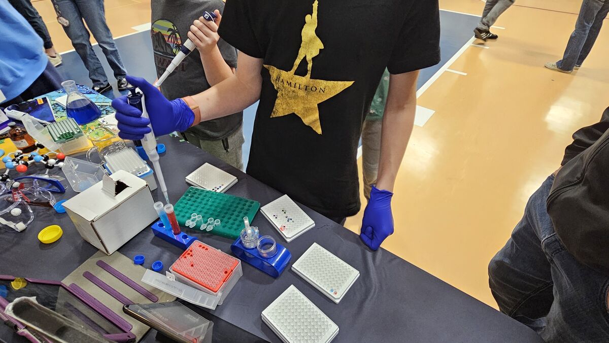 A person wearing blue gloves uses a pipette to transfer liquid into a test tube, surrounded by scientific equipment.