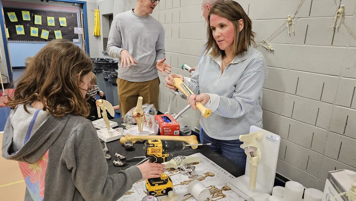 A young girl learns about the skeletal system with a teacher and a model of a human bone.