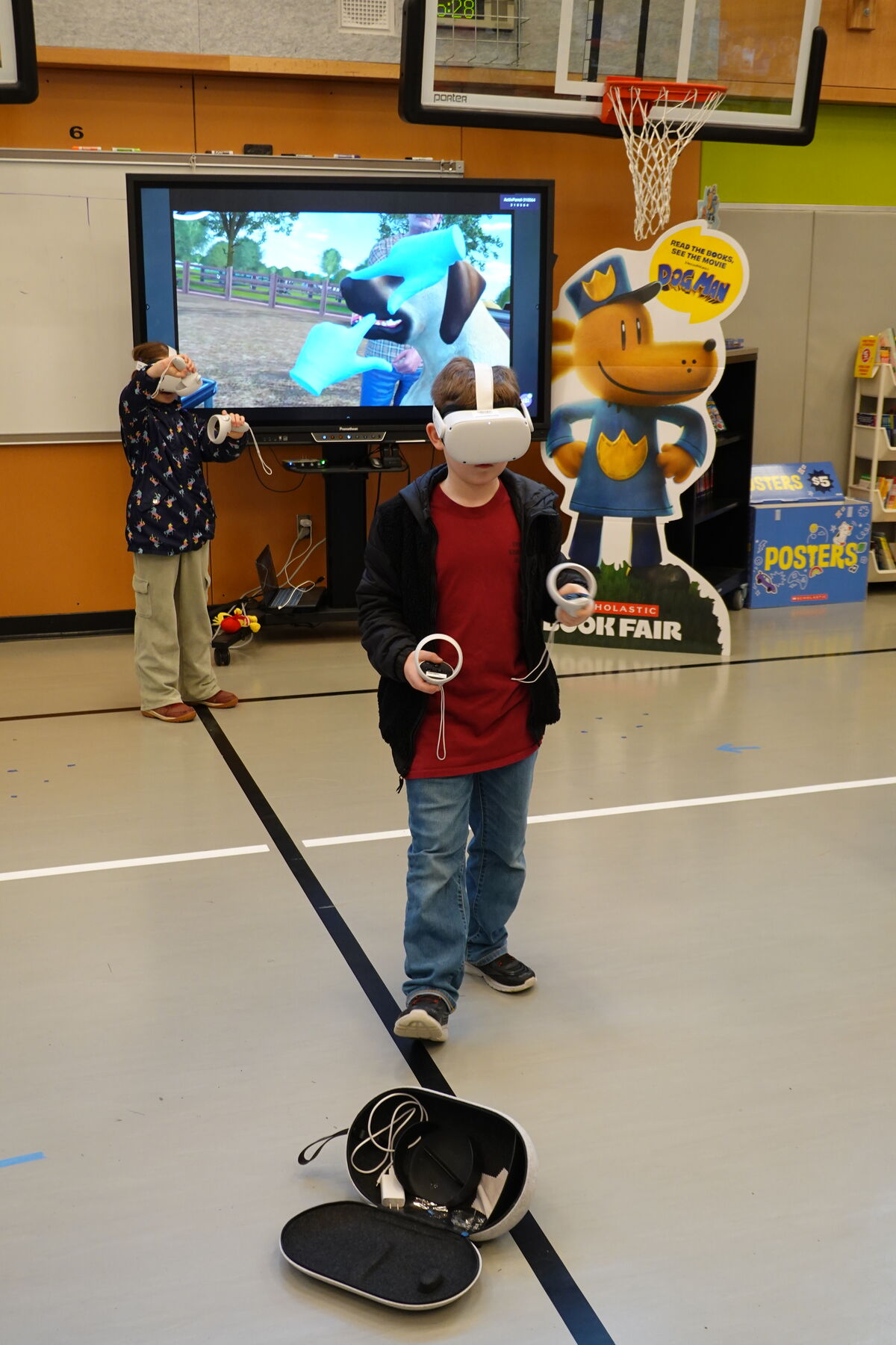 Two children use virtual reality headsets and controllers in a school gymnasium, with a large screen displaying a virtual world and a Dog Man cutout in the background.
