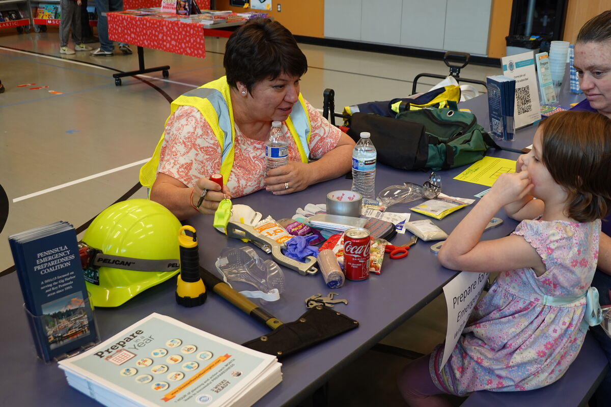 A woman in a yellow vest explains emergency preparedness to a young girl, with a table full of supplies between them.