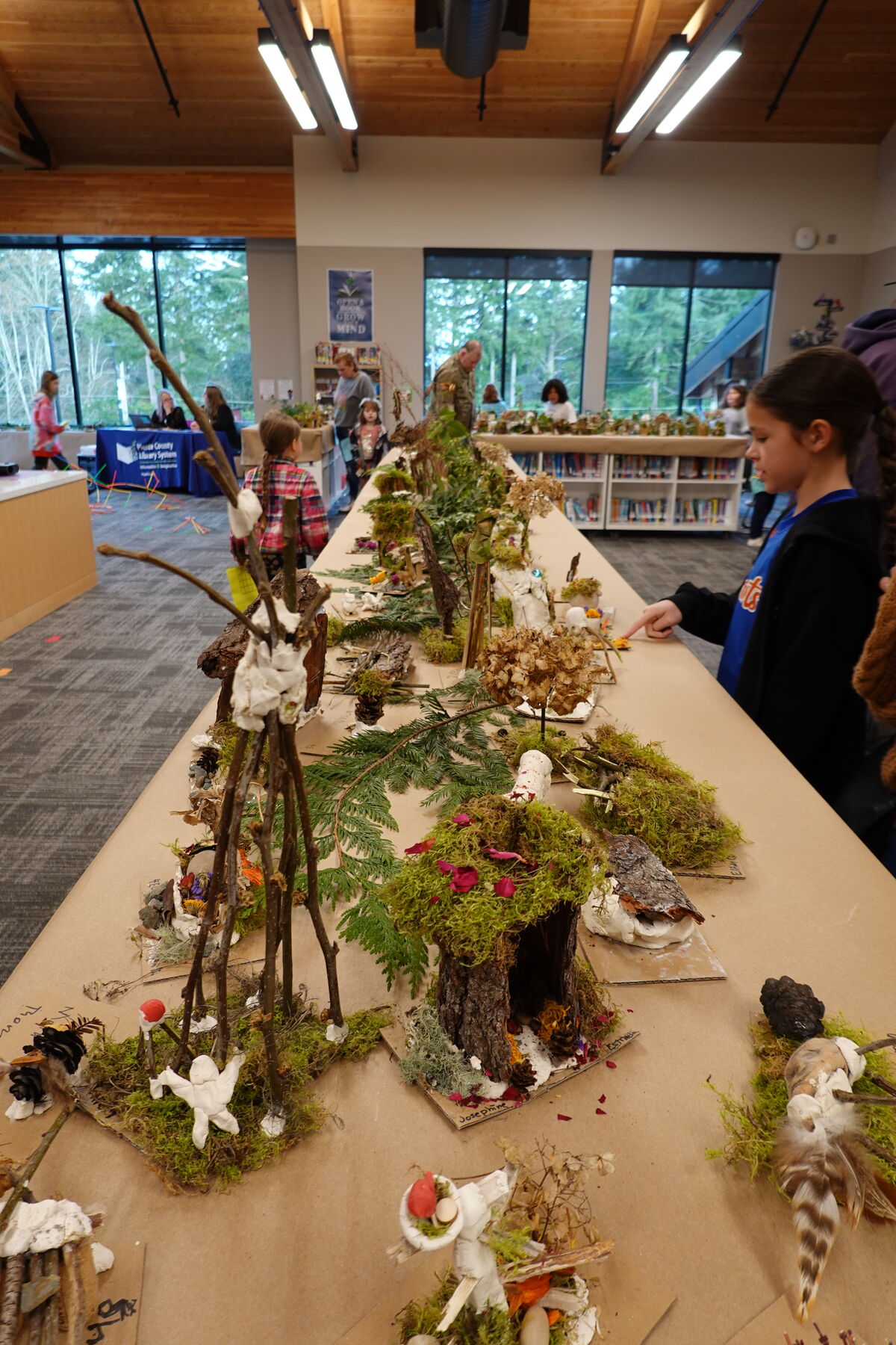 A long table displays intricate miniature fairy houses made from natural materials, with a young girl admiring the creations.