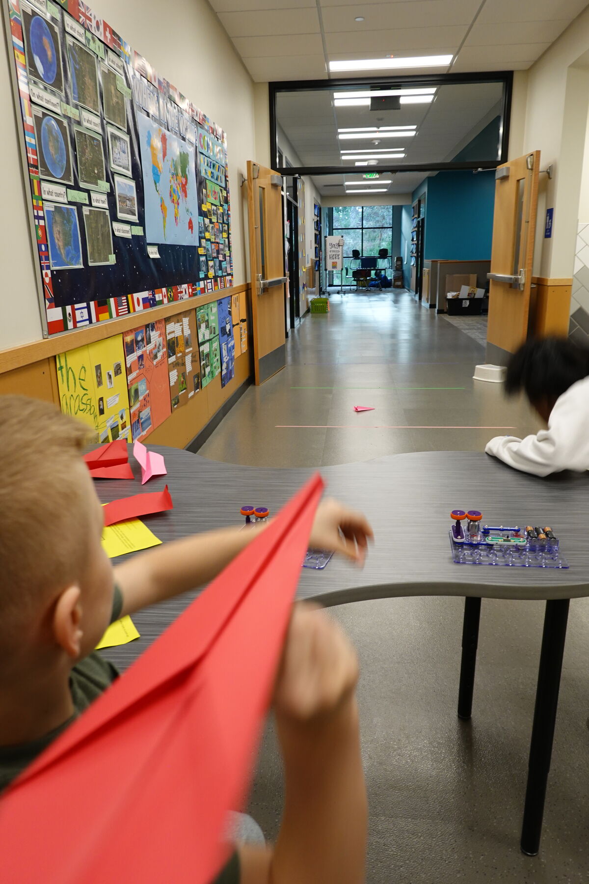 A child holds a red paper airplane in a school hallway, with a world map and other educational posters on the wall.