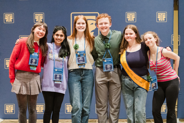 Six young people stand together, smiling and wearing name tags, in front of a blue wall with gold symbols.