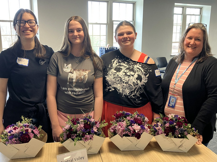 Four people stand in front of a table with floral arrangements, smiling for the camera.