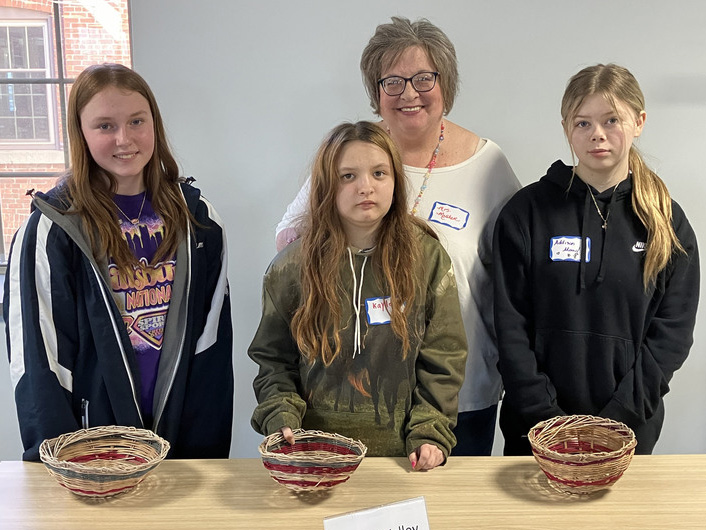 Four people, including a woman and three girls, stand in front of a table with three woven baskets. Each person has a name tag.