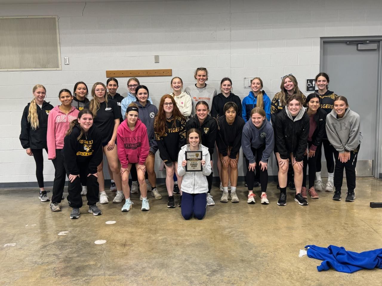 A group of young women stand together, smiling and holding a trophy. They are wearing athletic clothing and some have their school's name on their shirts.