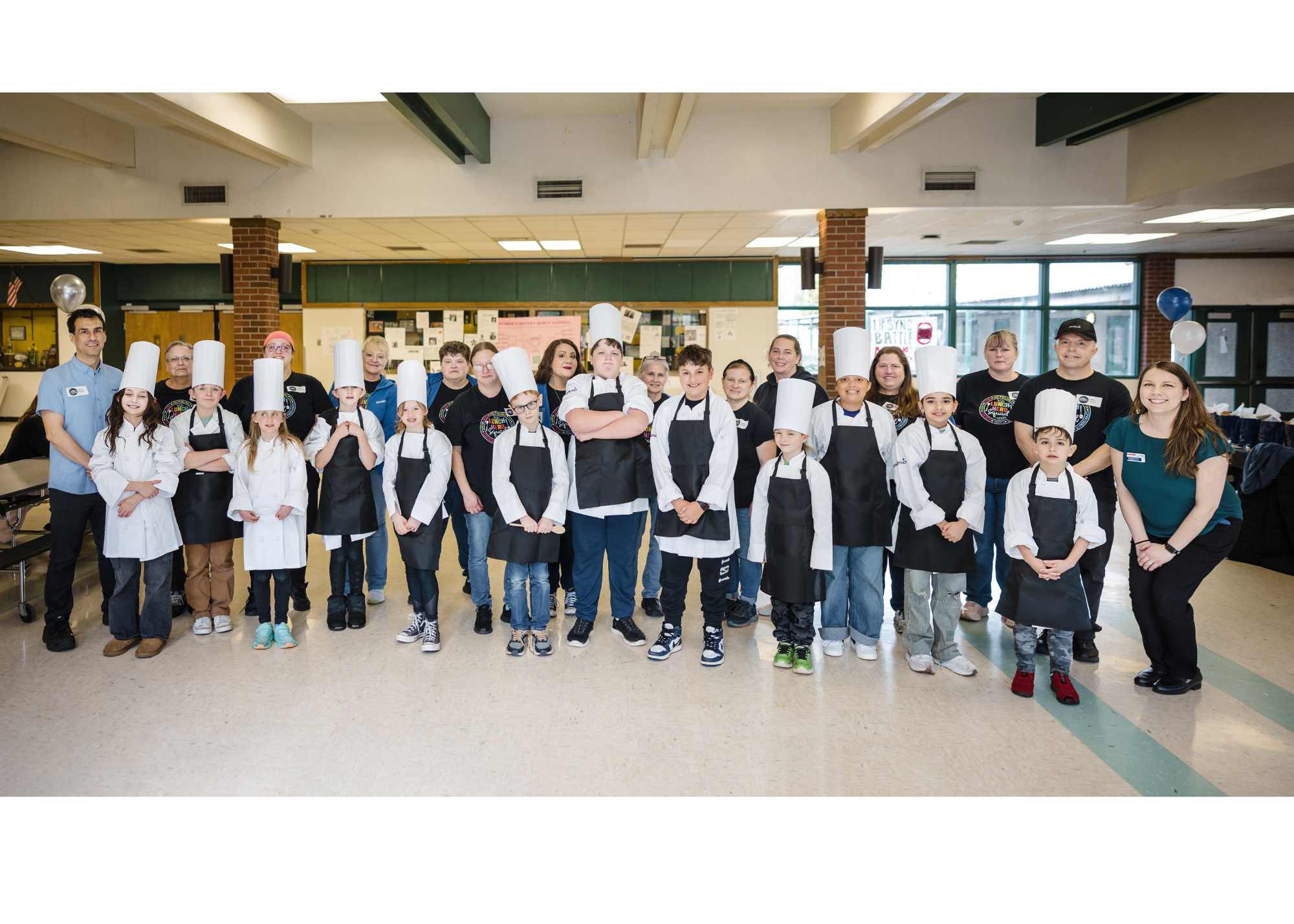 A group of children and adults stand together in a school hallway, wearing chef hats and aprons.