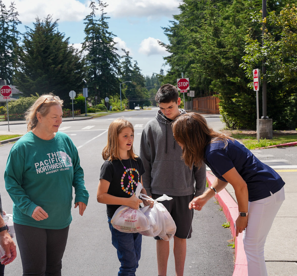 A group of people stand on a sidewalk, with a woman in a blue shirt talking to a young girl holding a plastic bag.