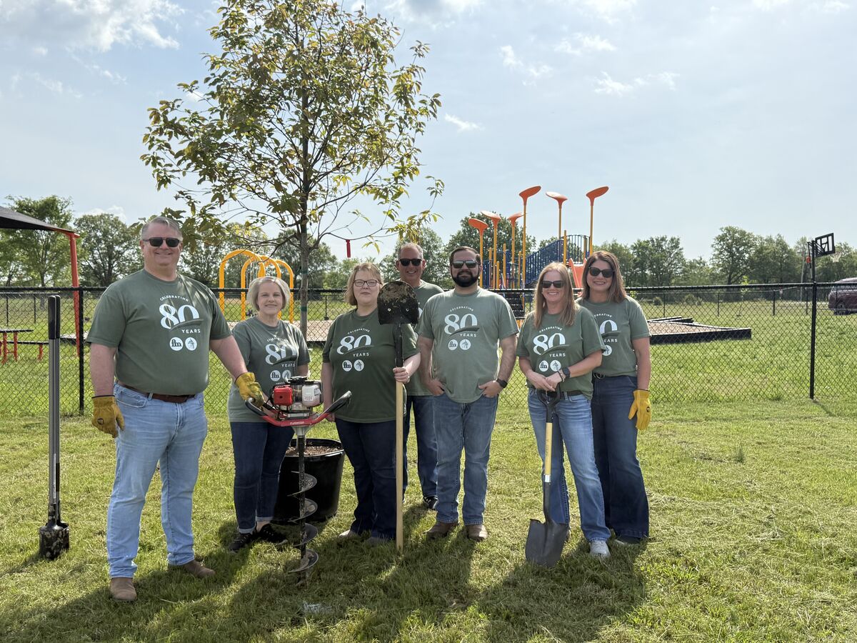A group of volunteers plant a tree on a playground, wearing shirts that say 'Celebrating 80 Years'.