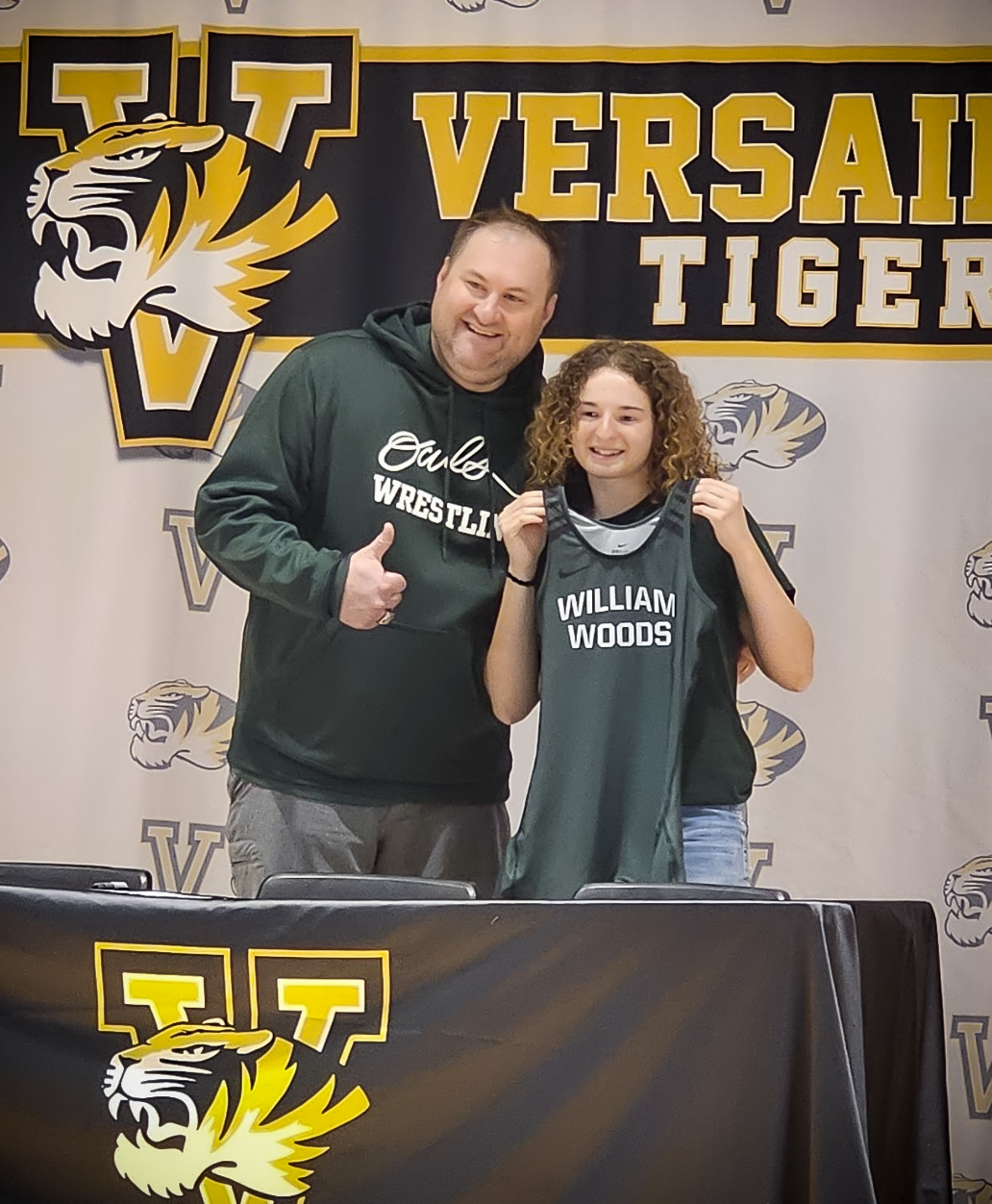 A young woman holds a green wrestling singlet with 'William Woods' written on it, standing with a man in front of a banner that reads 'Versailles Tiger'.