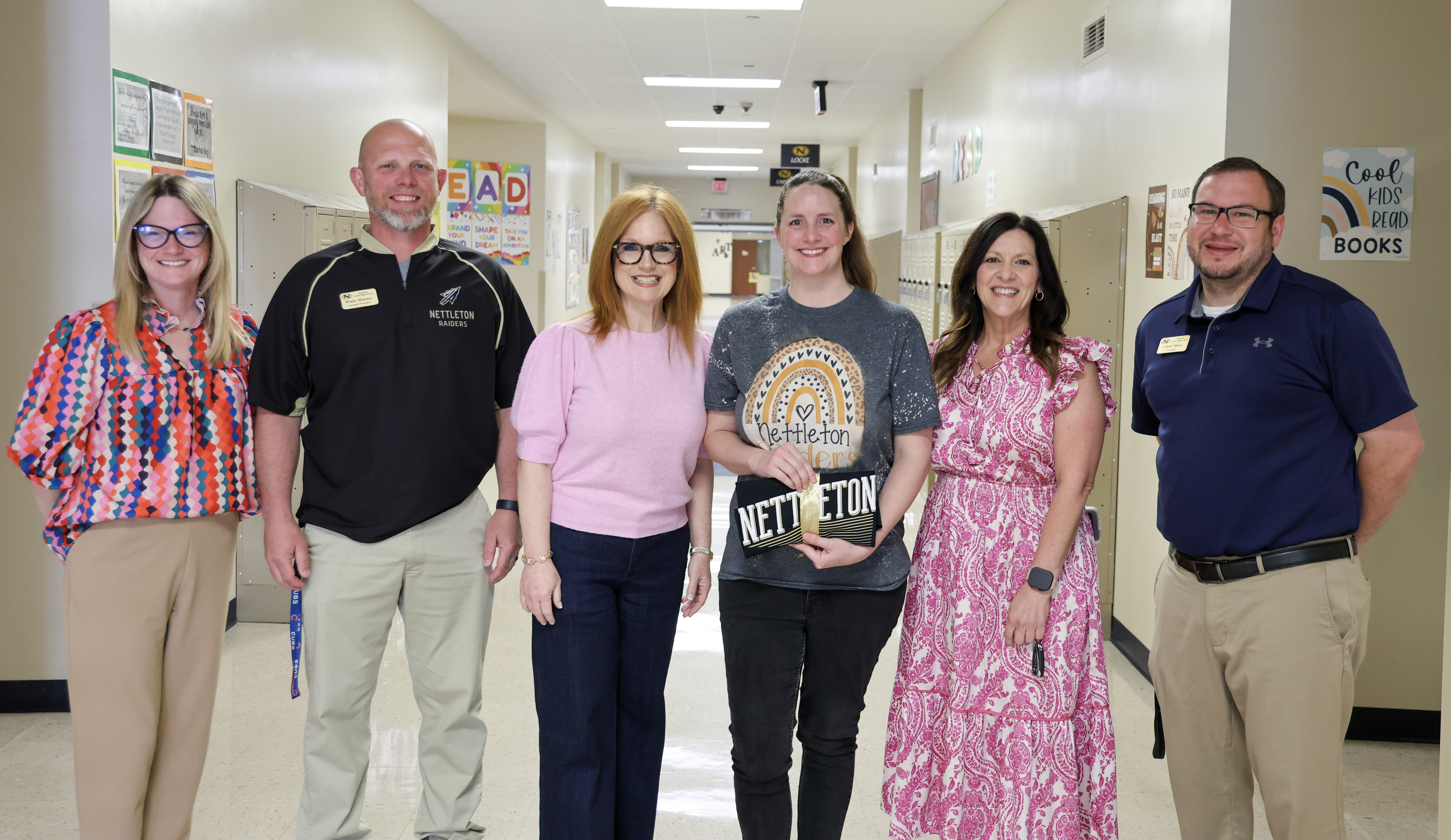 A group of five people stand in a school hallway, smiling for a photo. The person in the center is holding a black clutch with gold lettering that reads 'Nettleton'.