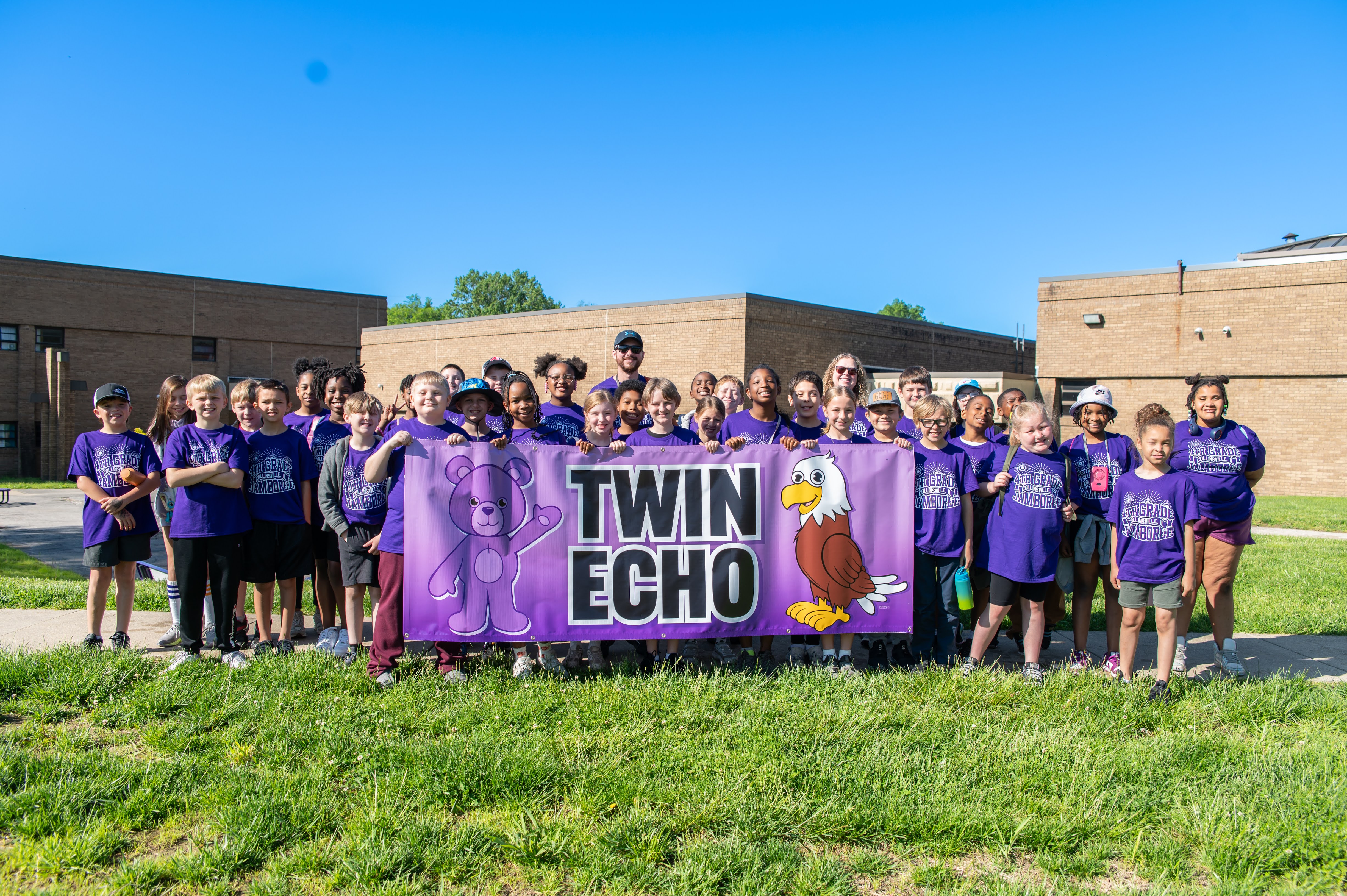 A group of children and adults stand together holding a banner that reads 'Twin Echo' with a teddy bear and eagle mascot.
