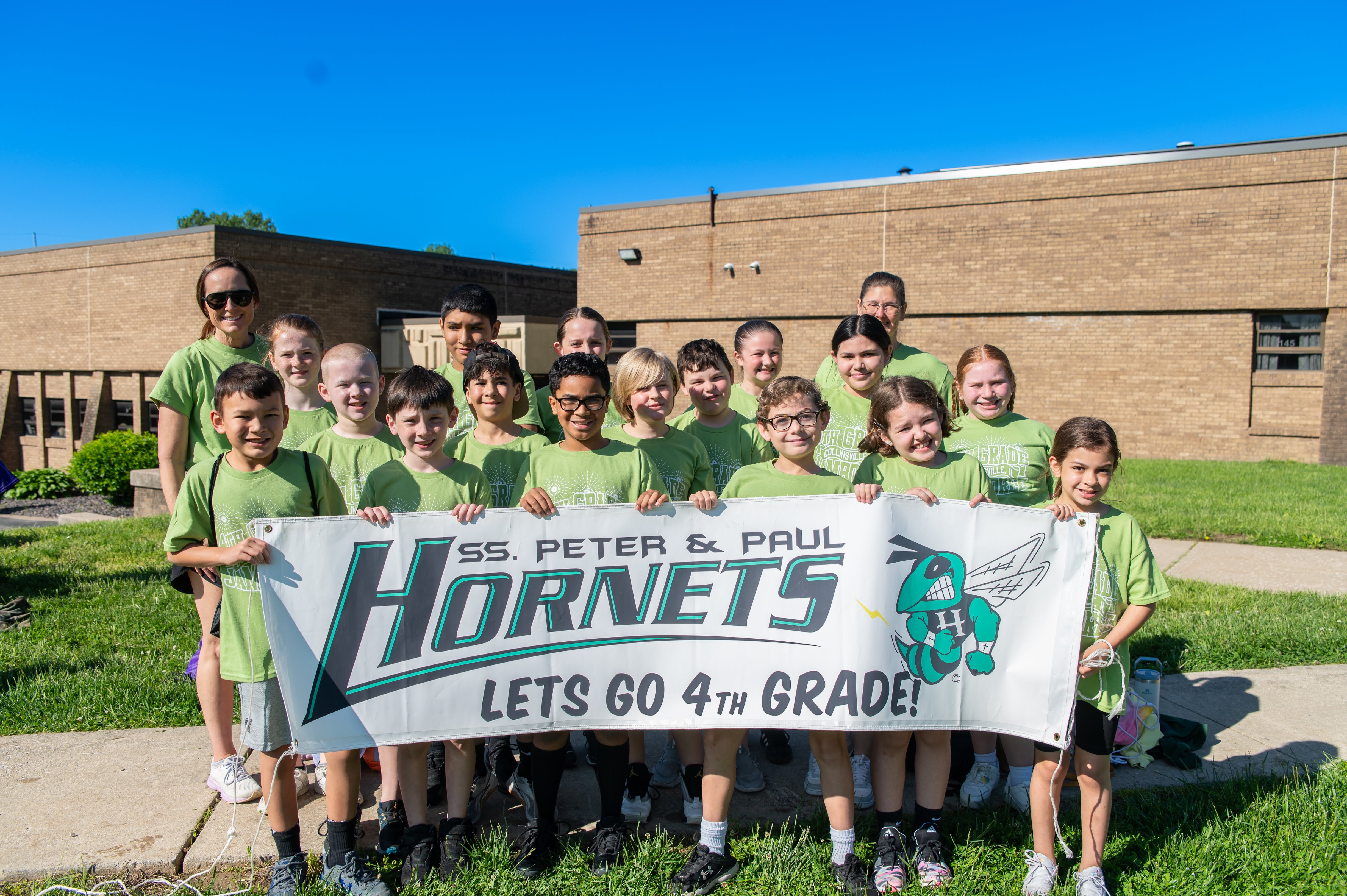 A group of children and their teacher pose for a photo in front of a school building, holding a banner that reads 'SS. Peter & Paul Hornets Lets Go 4th Grade!'