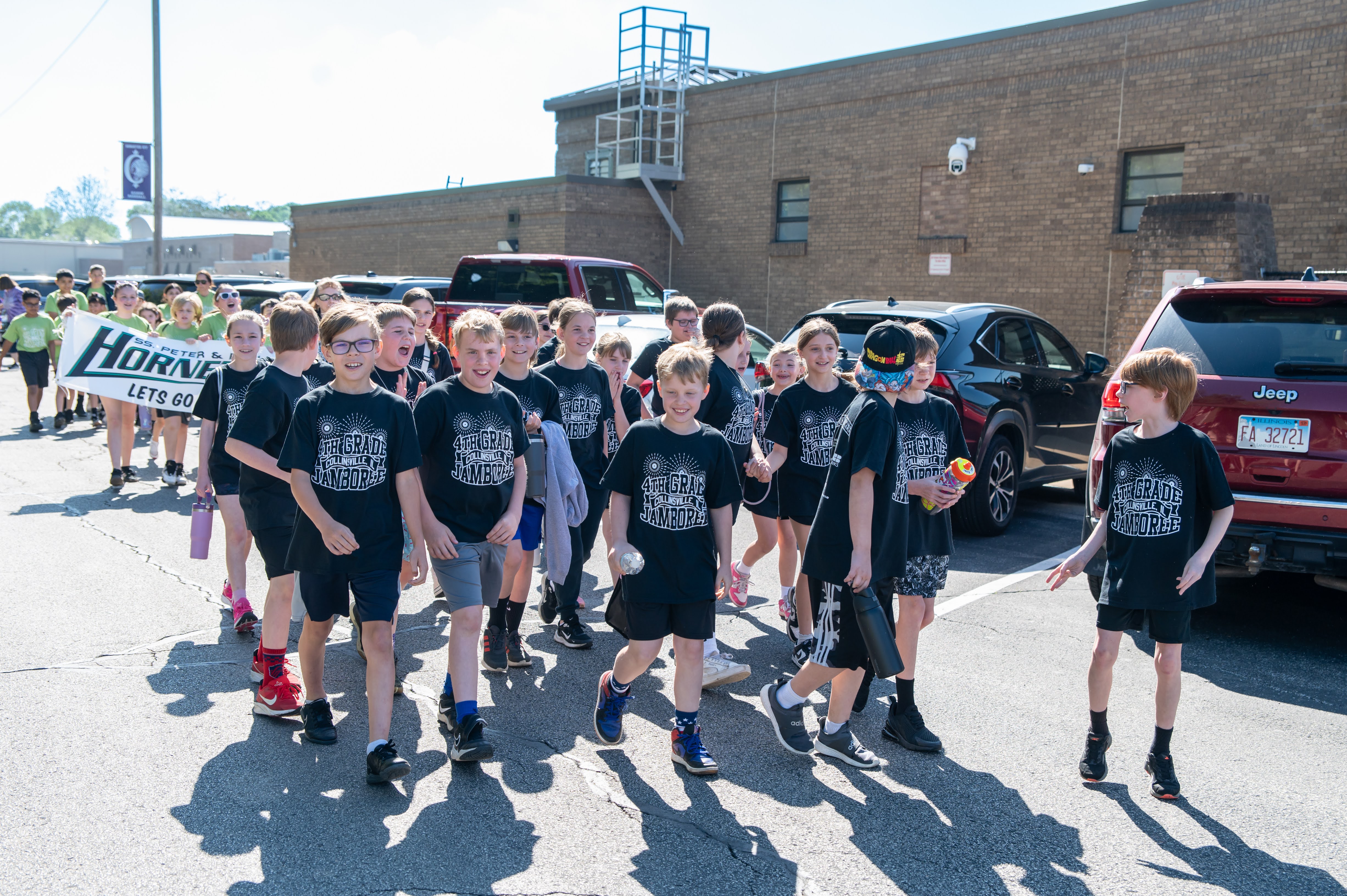 A group of children wearing black shirts with '4th Grade Collinsville Jamboree' printed on them walk in a line, some holding water bottles.