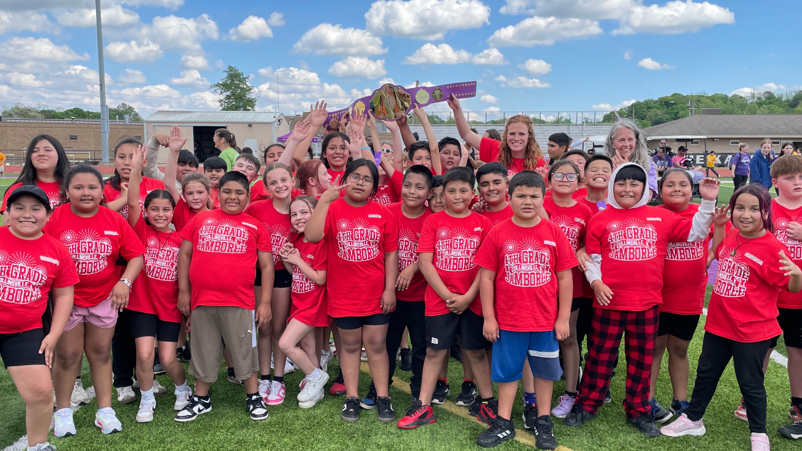 A group of children wearing red shirts with the text '4th Grade Collinsville Jamboree' hold up a trophy and smile for the camera.