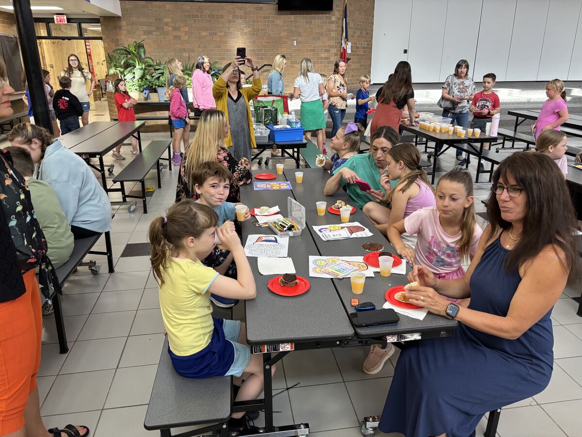 A group of people gather at tables, enjoying food and drinks in a school cafeteria.