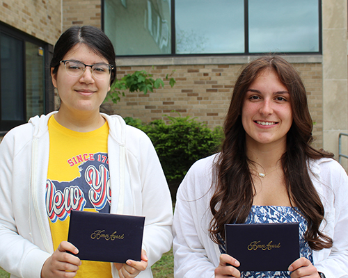 Two young women smile, holding certificates in front of a brick building.