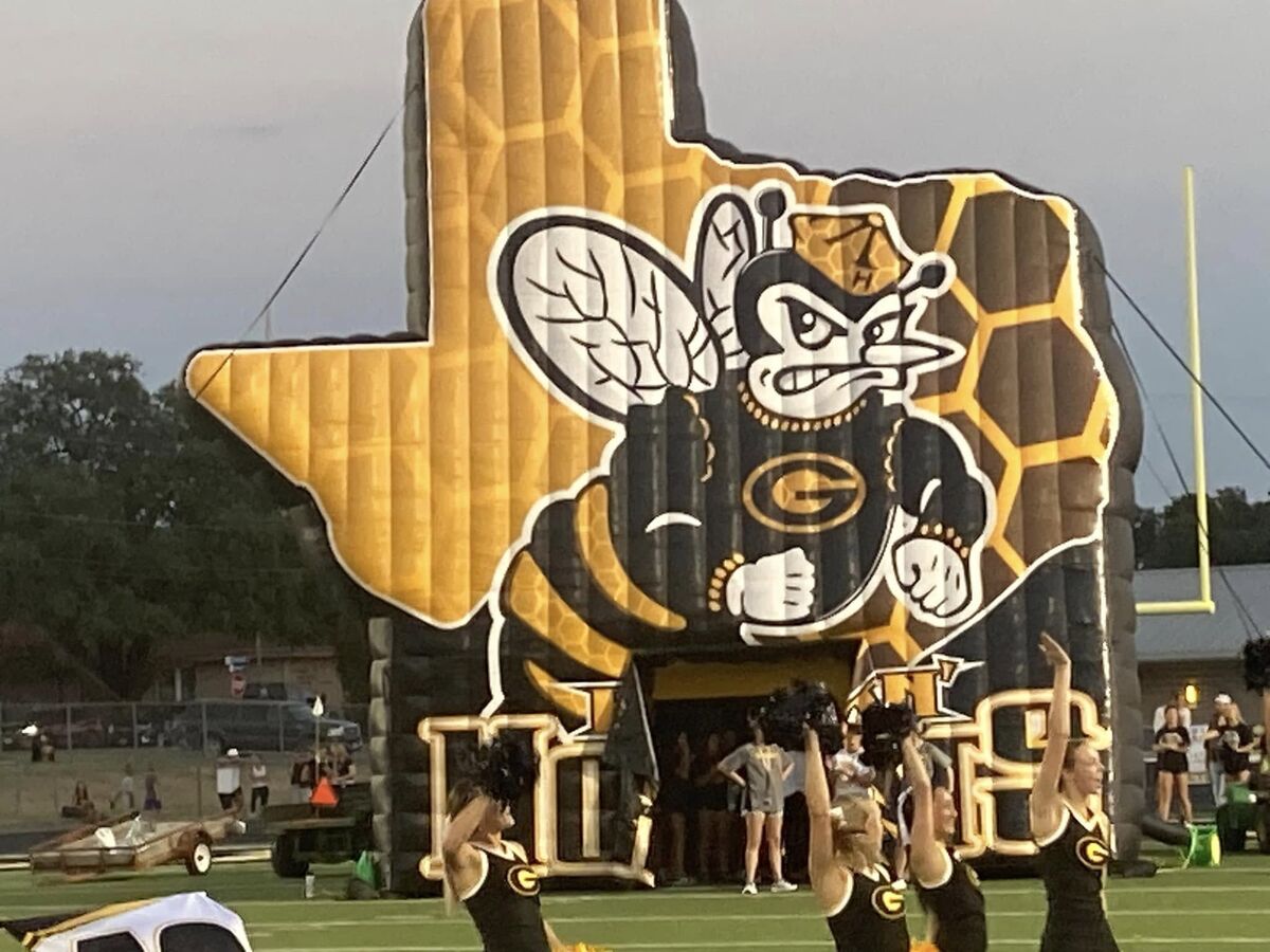 Cheerleaders perform in front of a large inflatable bee-themed tunnel at a football game.