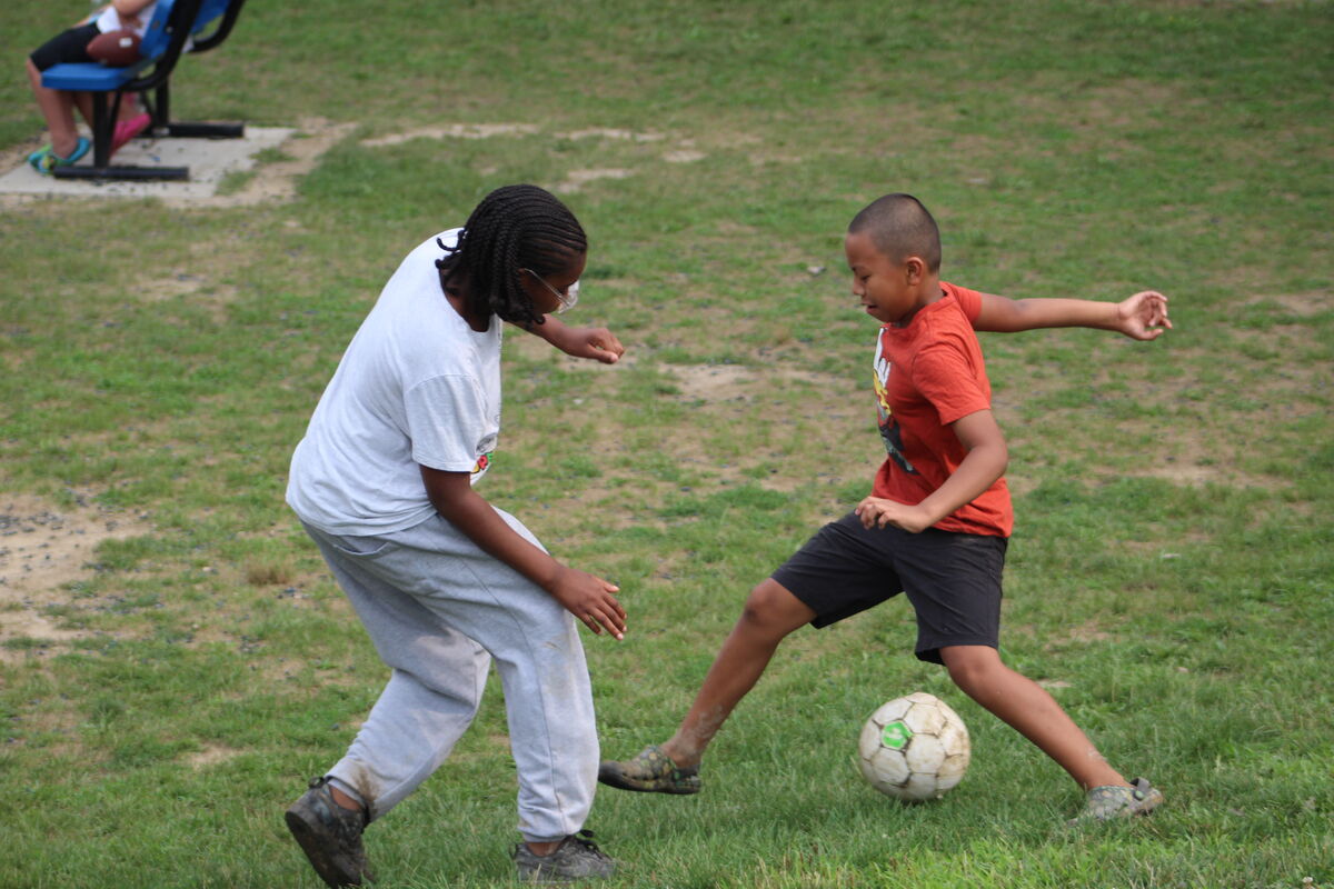 Two children play soccer on a grassy field, one kicking the ball.