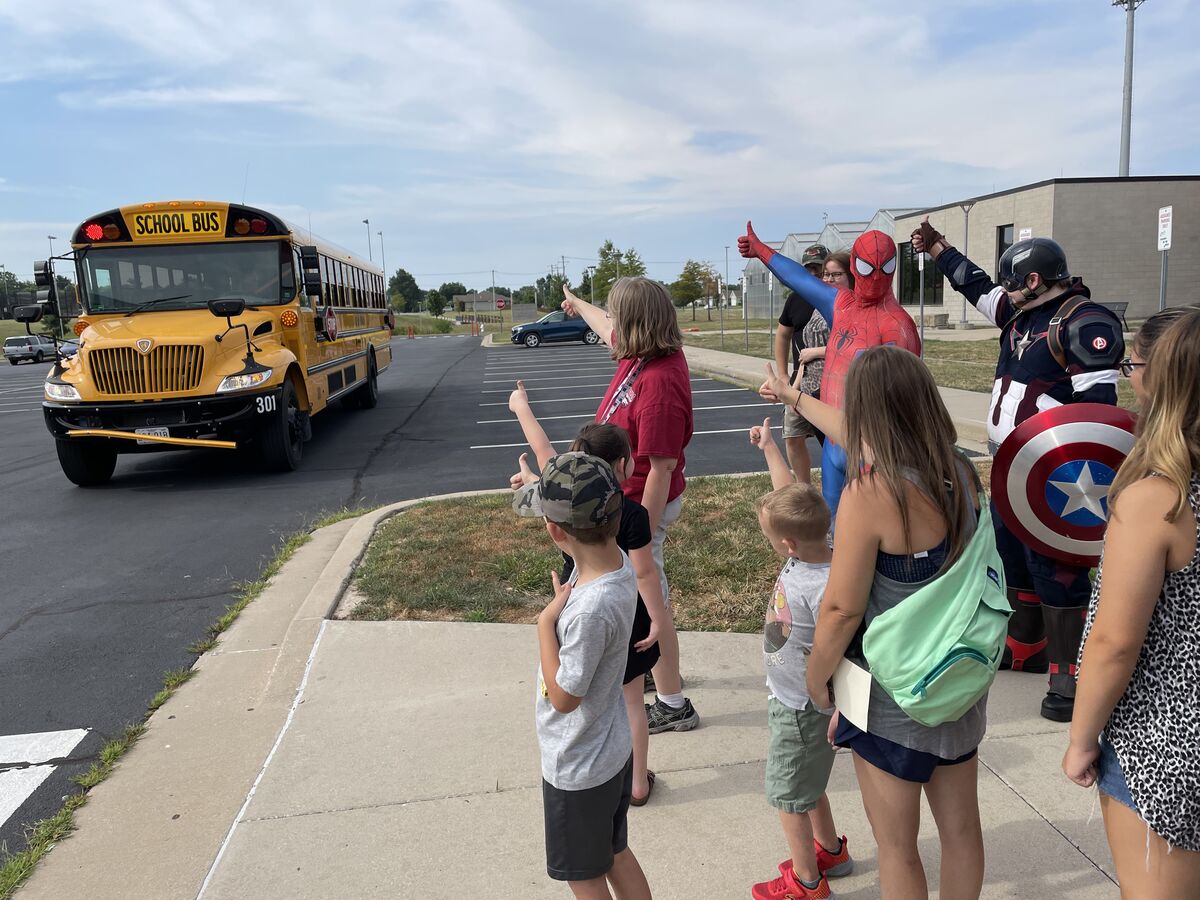 A group of children, parents, and costumed adults stand on the sidewalk waving as a school bus drives past.