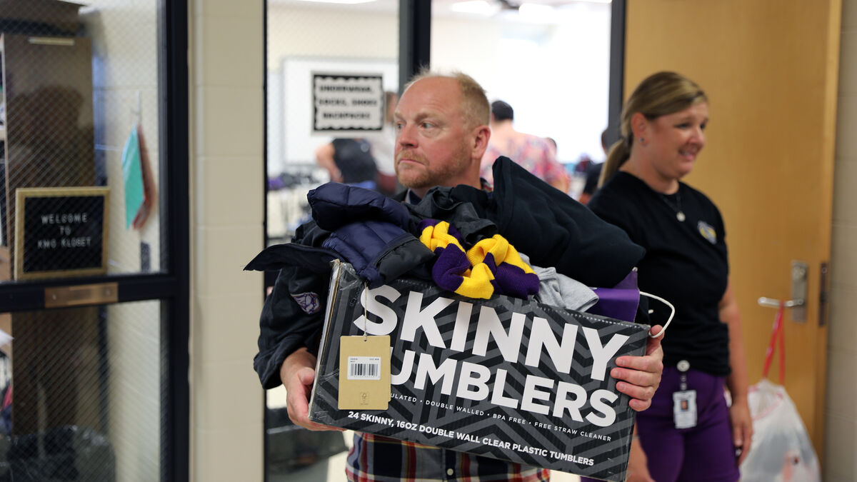 A man carries a box of 'Skinny Tumblers' through a hallway, followed by a woman.